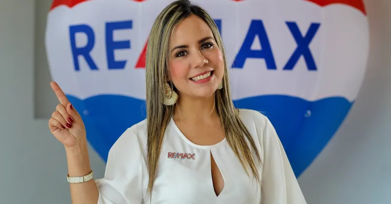 Smiling real estate agent in white top pointing with RE/MAX balloon in the background.